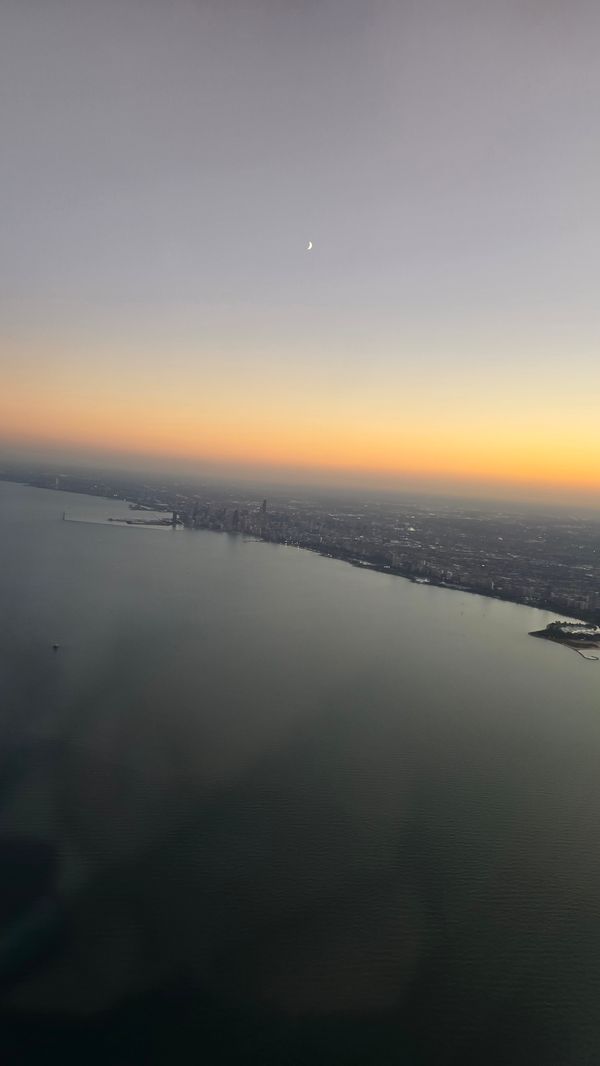 A shot of Chicago from the plane, with the shot dominated by Lake Michigan. It is sunsent, and the crescent moon is visible over the city.