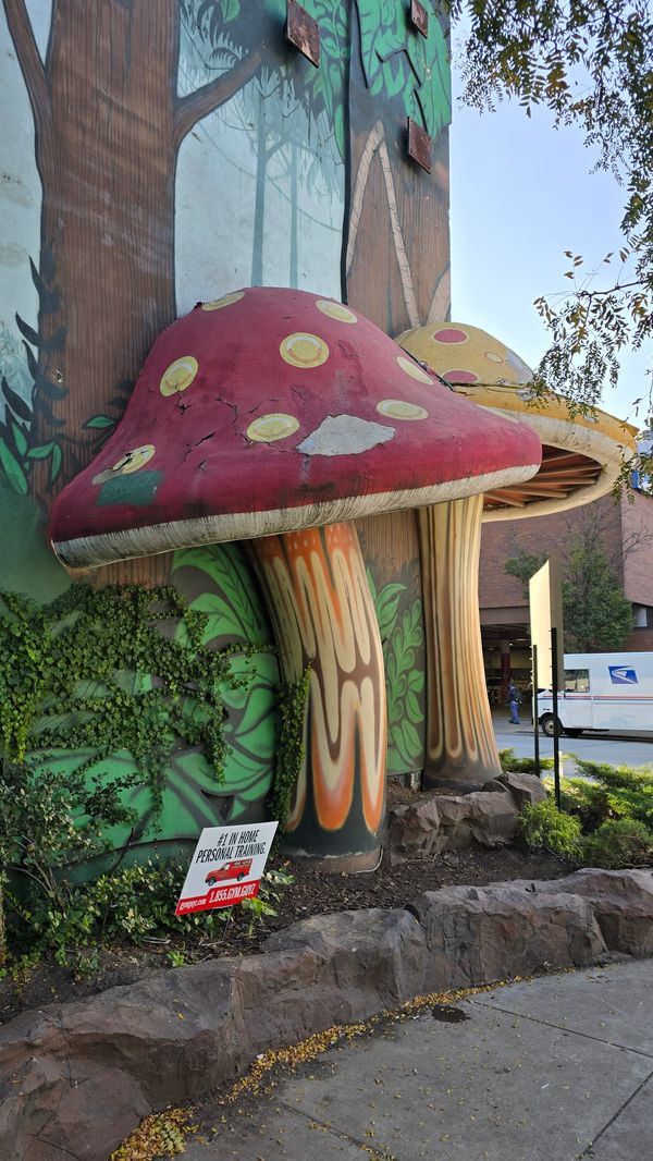 A large, fake, red mushroom attached to a building that is covered in a mural depicting a rainforest. A bit of a yellow mushroom in visible behind the red one.