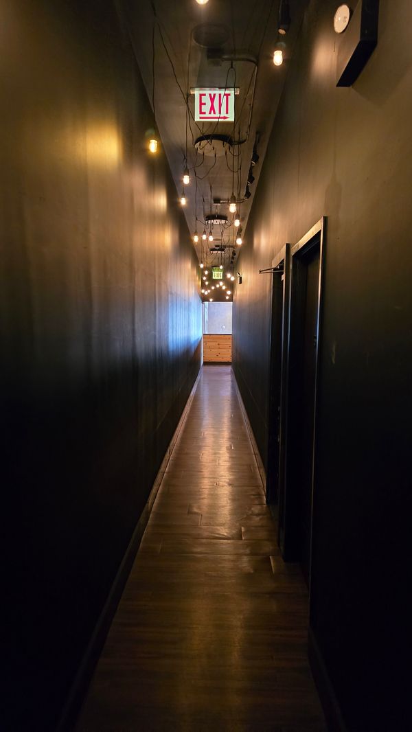 A long, narrow, dark hallway in a restaurant (Pilsen Yards). It is illuminated by bare bulbs on a string lights, and a pair of EXIT signs.