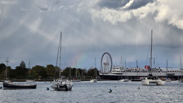 A photo of the Navy Pier Ferris wheel in the distance , with several boats on Lake Michigan in the foreground. Right next to the Ferris Wheel is a rainbow.