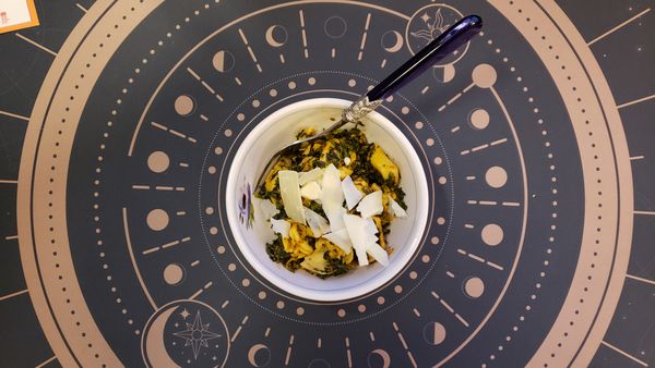 A photo of a bowl of tortellini and spinach in a tomato sauce. The bowl is set on a deskmat that depicts a sort of magic circle with iconography of the sun, the moon, and stars.