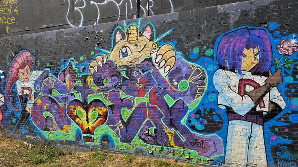 A graffiti wall, with a tag styled after the Pokemon Arbok. Around the tag are Jessie, James, and Meowth.