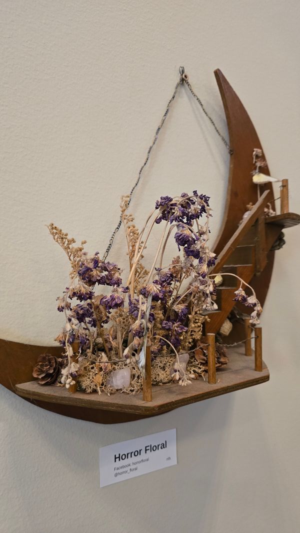 A wooden wall hanging, shaped like the moon. Attached to the moon is a staircase, and at the foot of the stairs are a bunch of white and purple plants. The piece is labeled 'HORROR FLORAL Facebook: horrorfloral @horrorfloral'