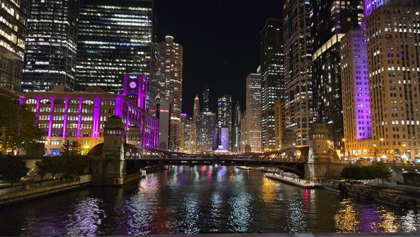 A photo over the Chicago River at night, showing a bridge, and buildings on both sides lit up with purple lights.