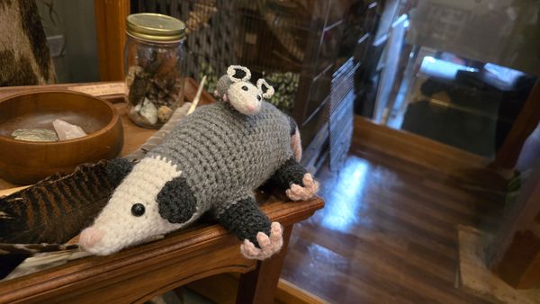 Two crocheted stuffed animal opossums, one is much smaller and riding on the back of the larger one.