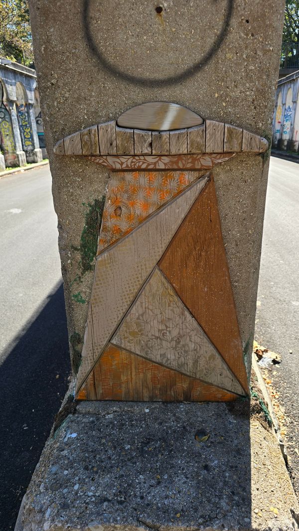 A wooden carving of a UFO beaming up something, with the pieces of wood seemingly coming from several different pieces of scrap wood. The whole piece is attached to a concrete pillar.