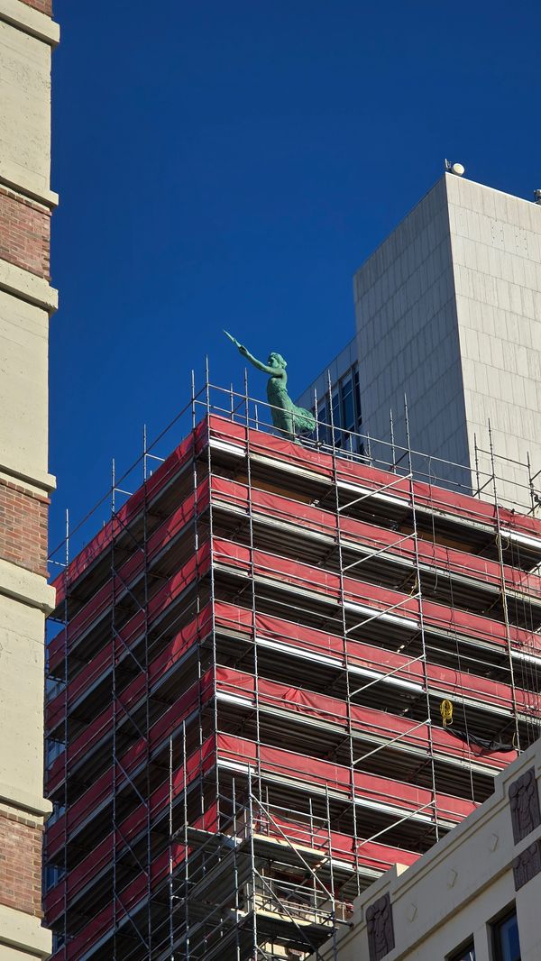 Scaffolding around a building topped with a green statue pointing to the left of frame.