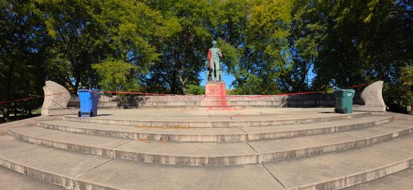 A photo of a status of Abraham Lincoln, which is vandalized and roped off with red tape. The statue has been splashed with red paint, and on the pedestal is written 'Lincoln was an Executioner' A long bench in the background says 'May all empires fall, from Turtle Island to Palestine'