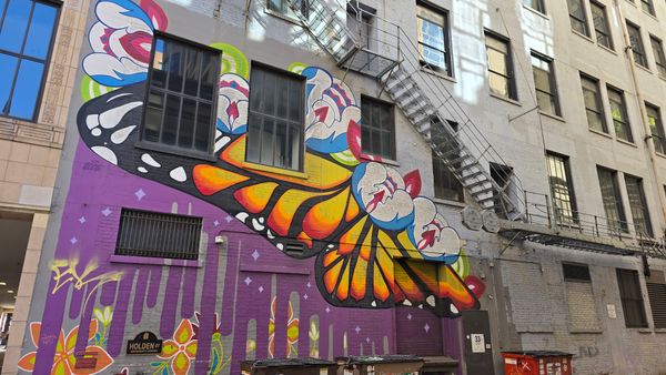 A mural on a building, underneath some fire escape stairs. The mural is abstract, showing a monarch butterfly wing coming out of several white, red, and pink blobs. The background is a dripping purple on a gray building, with colorful flowers painted near the ground.