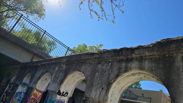 A concrete footpath from below, with several  concrete arches in the foreground. The whole shot is at an angle showing the intersection of the arches and the footpath. The columns of the arches have various bits of graffiti.