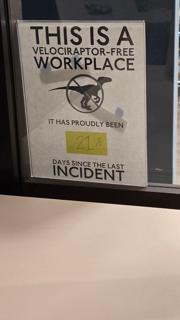 A taped-up sign on a window: 'This is a Velociraptor-free Workplace' with 'It has proudly been 21 days since the last incident' The '21' is written in on a post-it note, which also has a sad face drawn on it.