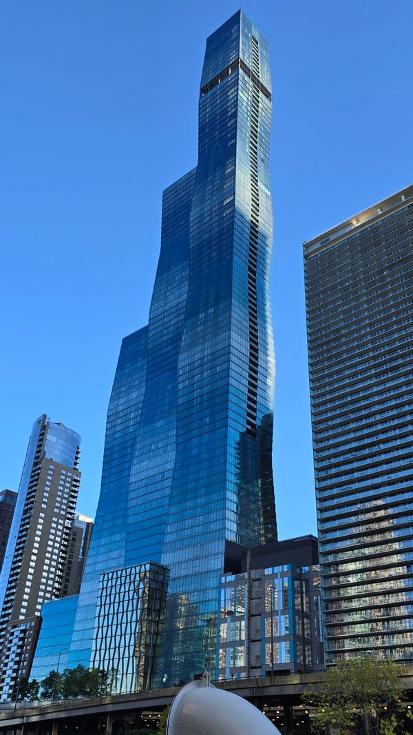 A tall glass building with 3 sections, each taller than the previous. Each section varies in width as they reach to the sky to create a wavy pattern.