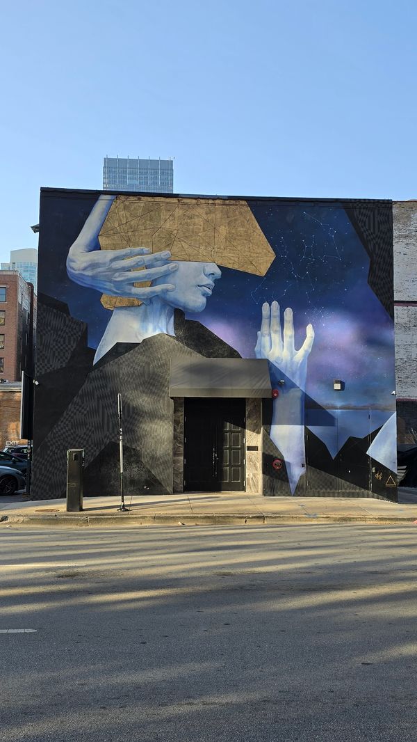 A mural taking up the entire side of a building, depicting a person wearing a gold, angular mask, with one arm wrapped around their head and one hand raised in front of them. The background shows a starry view with some constellations laid out.